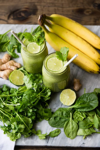 two mason jars filled with green smoothie surrounded by cilantro ripe bananas