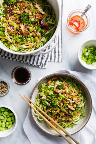 big bowl of zucchini pasta next to a shallow bowl with a helping of zucchini pasta with chopsticks resting on top surrounded by garnishes