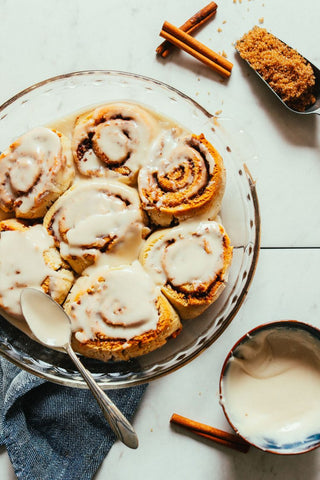 bowl with multiple cinnamon buns in the process of being iced with cinnamon stick nearby