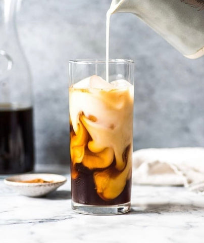 milk poured into tall glass of cold coffee with ice on a table with a jug in the background