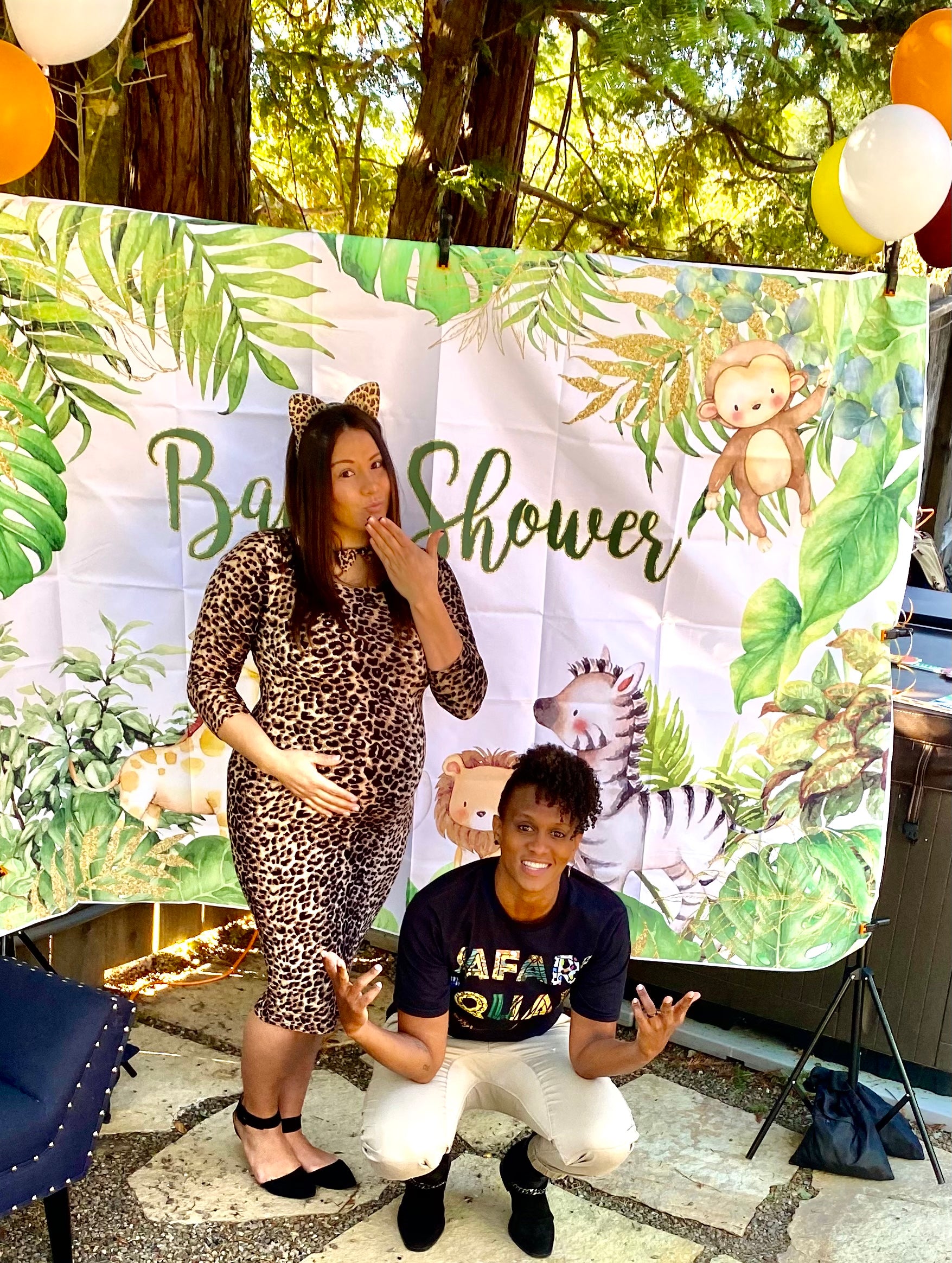 Pregnant mom in leopard print dress standing, and wife crouching next to her in black shirt and white pants at zoo themed Mosie Baby shower..