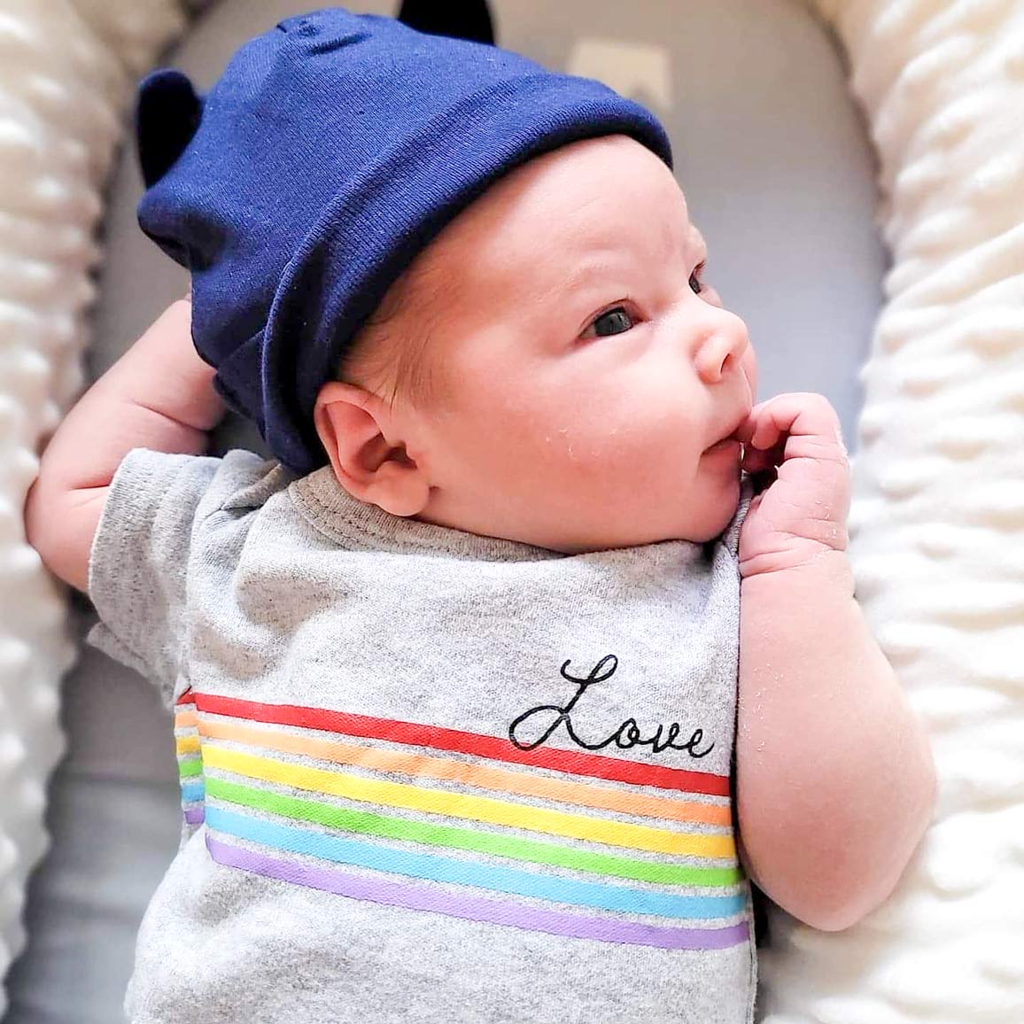 Baby boy in blue beanie hat and gray shirt with rainbow that says "love" looks up and to the right in his baby bed.