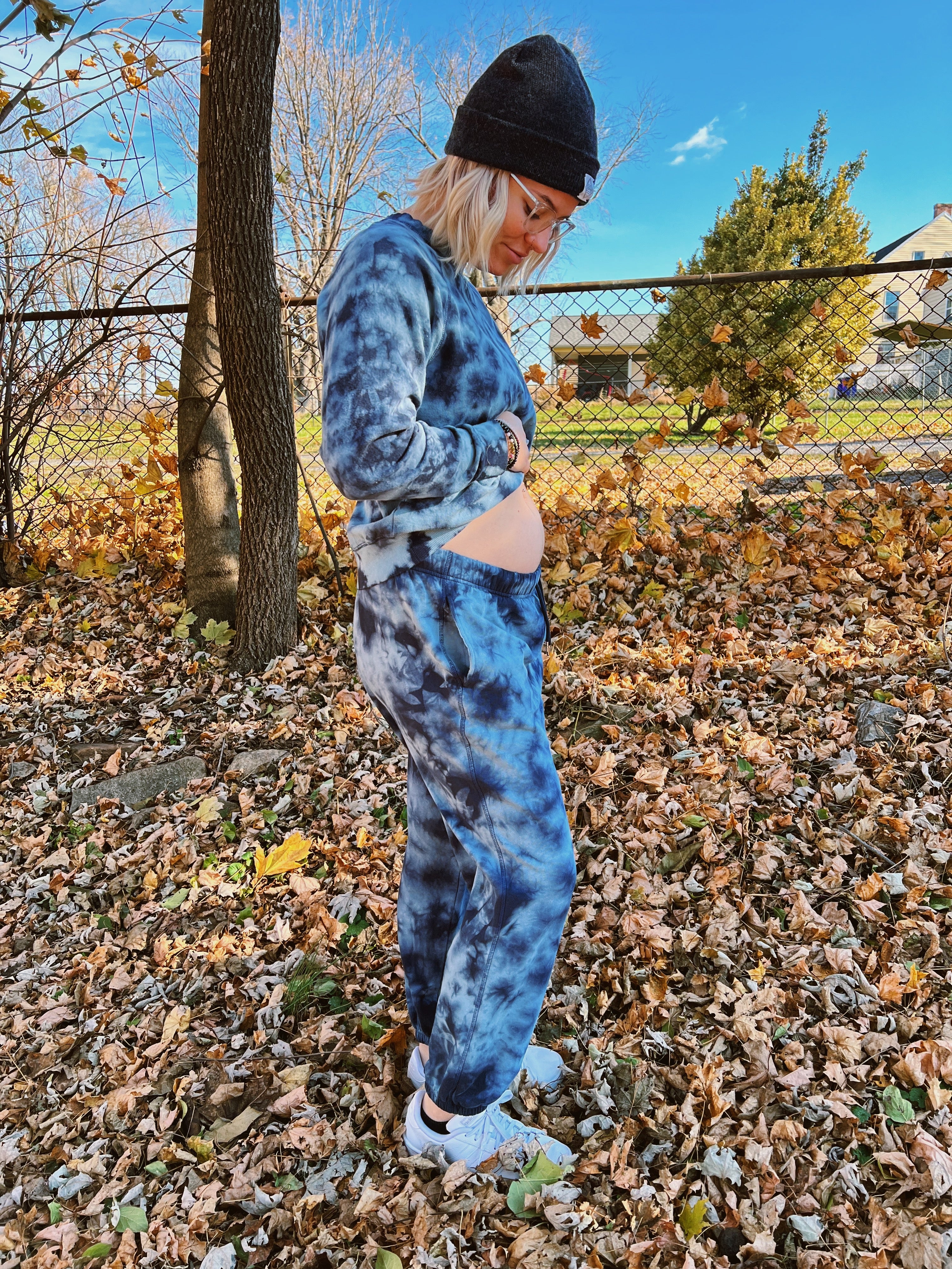 Pregnant woman in blue sweat shirt and matching sweatpants holds her exposed belly on a fall day