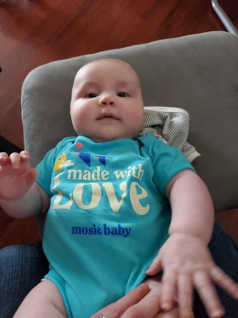 Mosie Baby in teal onesie laying down and reaching for camera