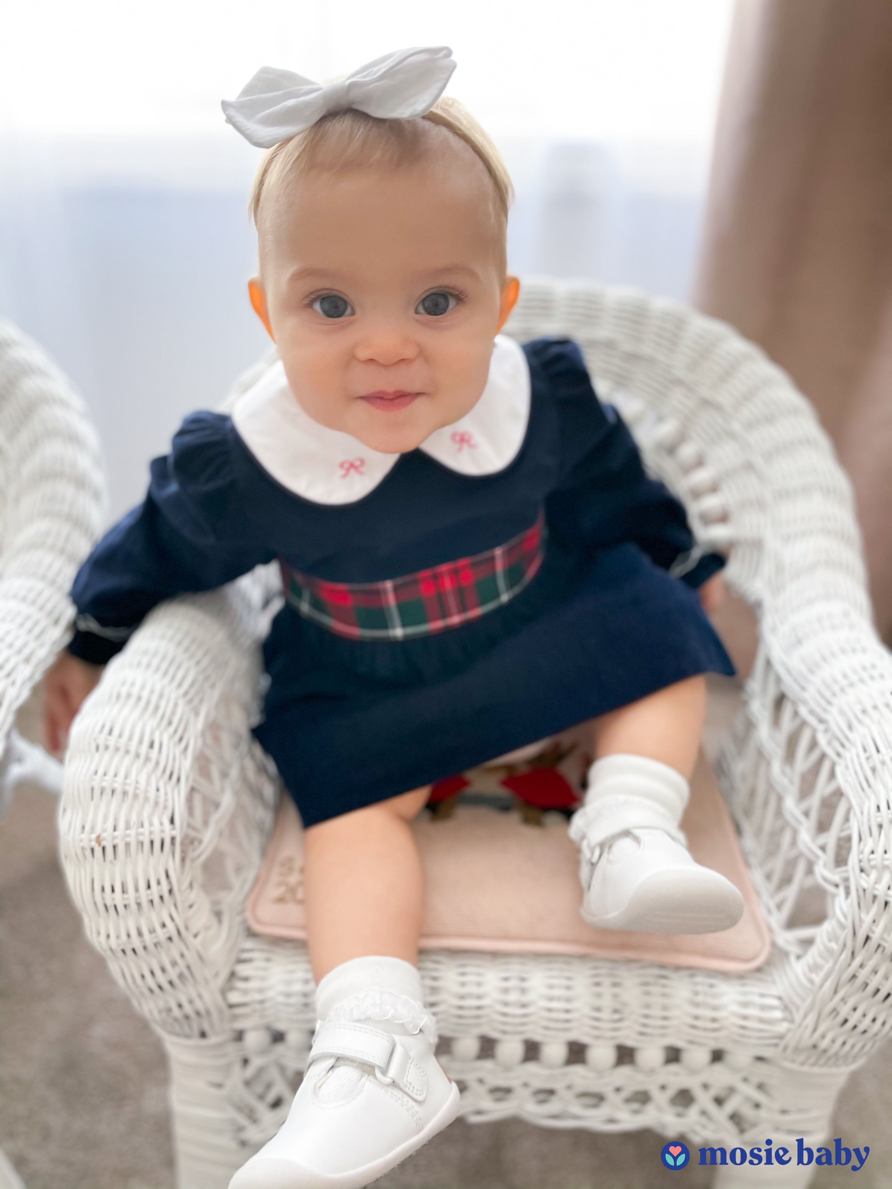 Mosie baby sitting in a white chair with a blue dress.