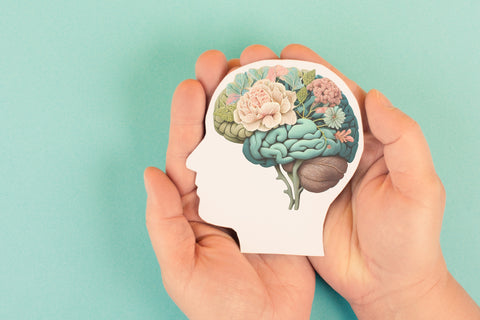 Hands holding paper cut out of a human head side profile with a brain of wellness flowers, rocks, plants, nature