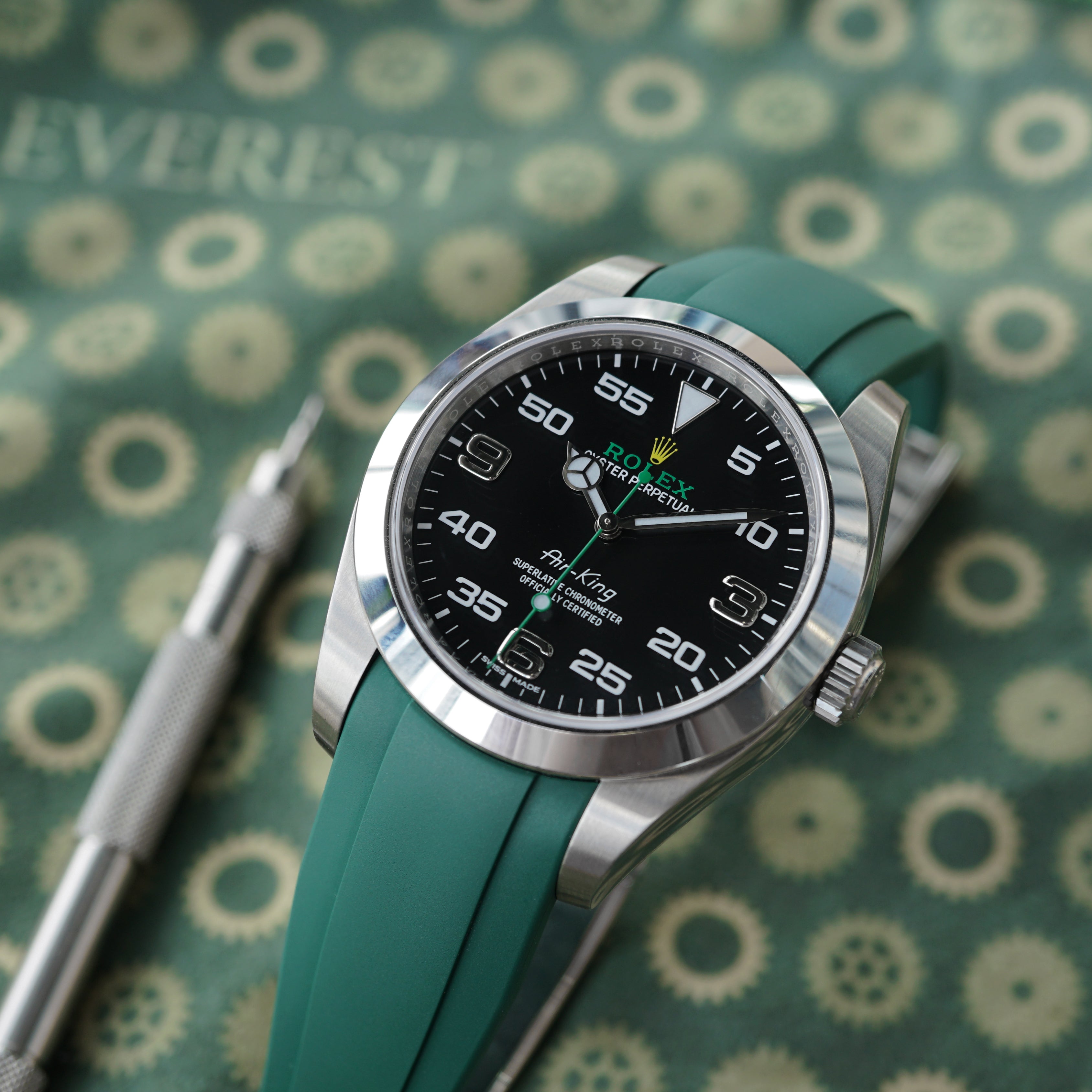 a rolex airking watch with a green rubber strap on it next to a metal spring bar tool