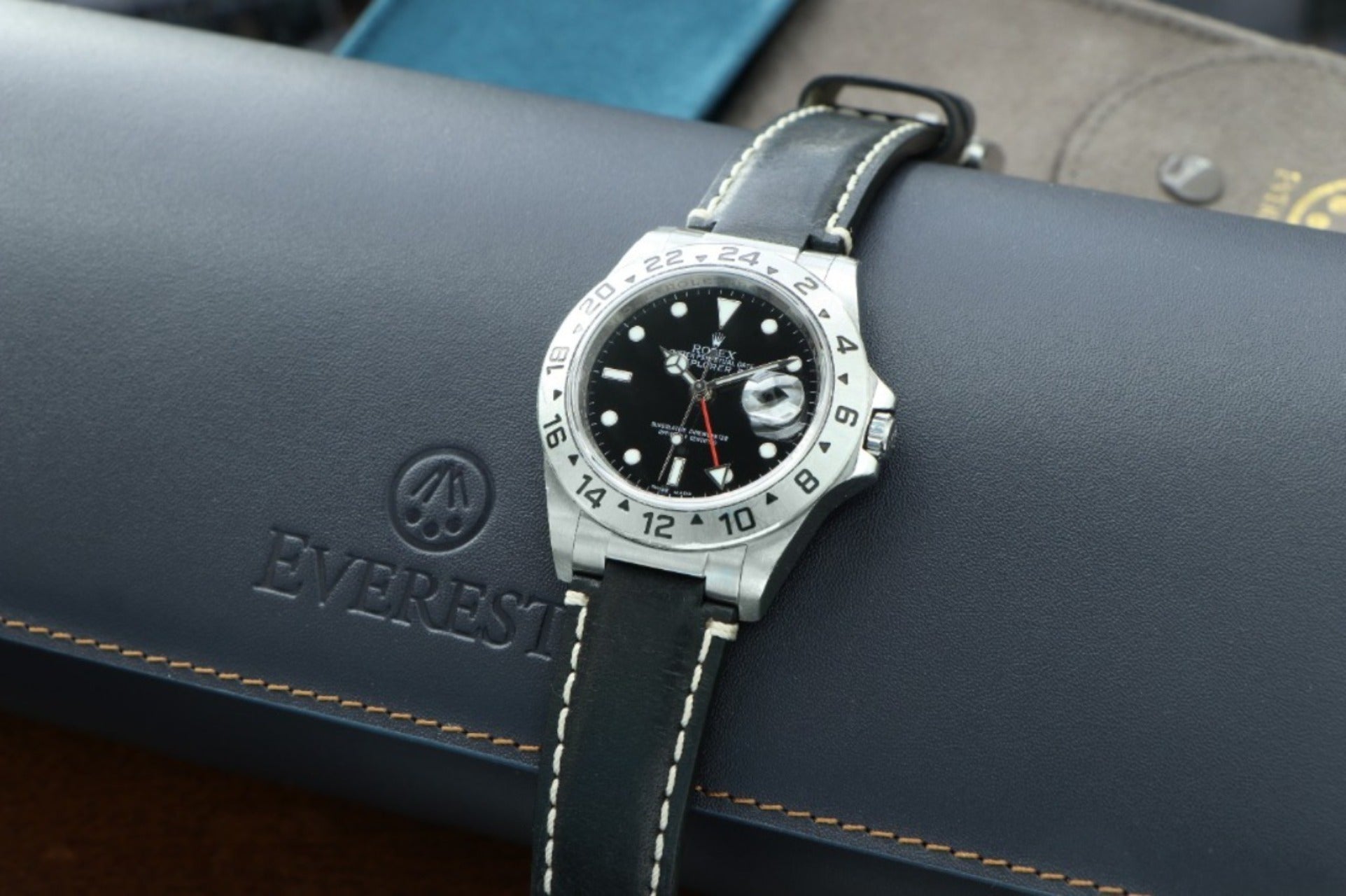 rolex explorer ii watch on a black leather strap with white stitching sitting on top of a blue leather watch roll