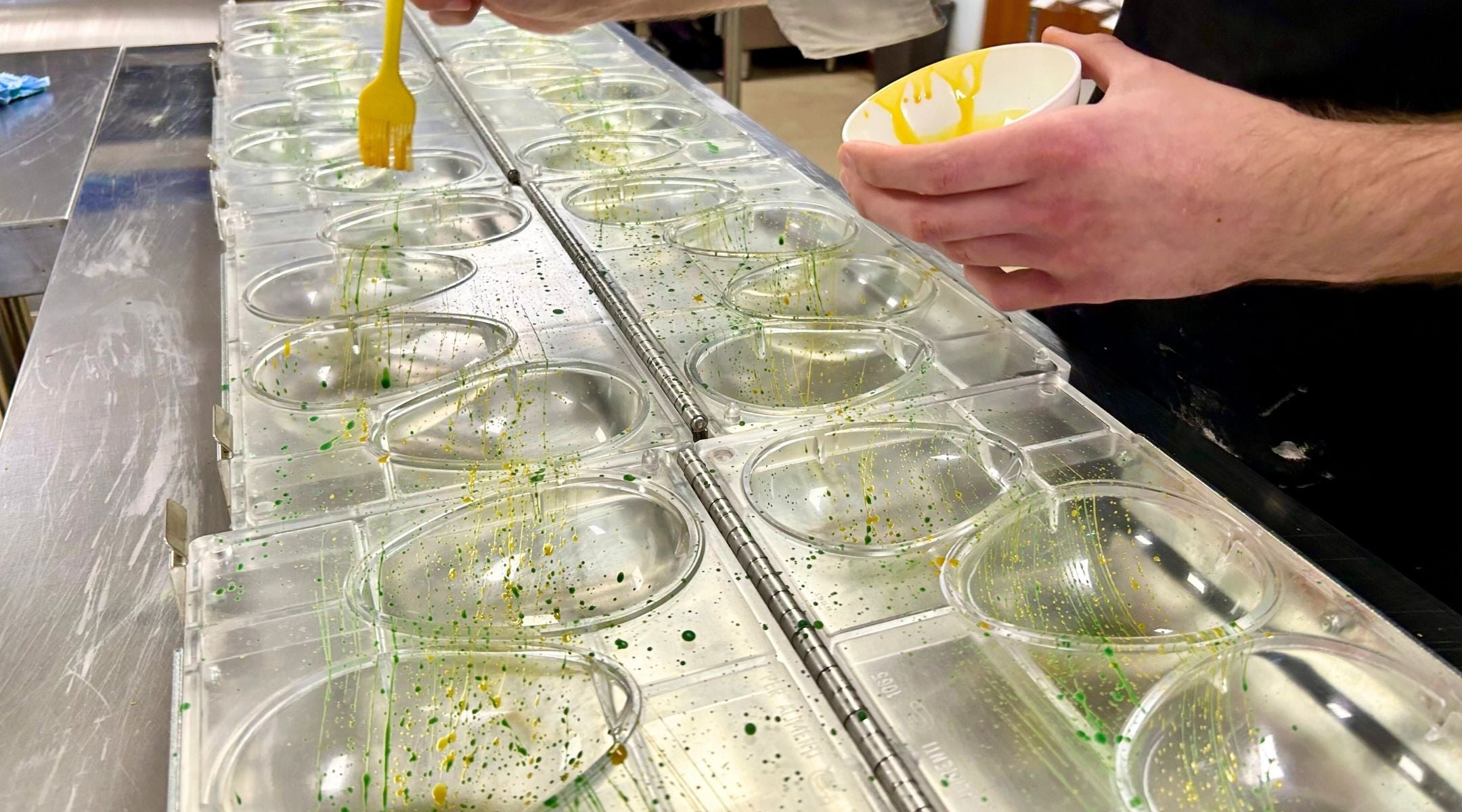A chocolatier hand-decorating a series of Easter egg moulds with yellow and green cocoa butter speckles and splatter