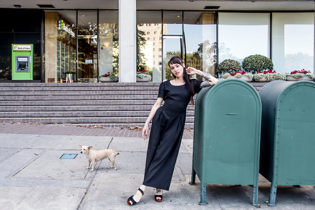 woman leaning on mail box with a black high waisted jump suit and little dog 