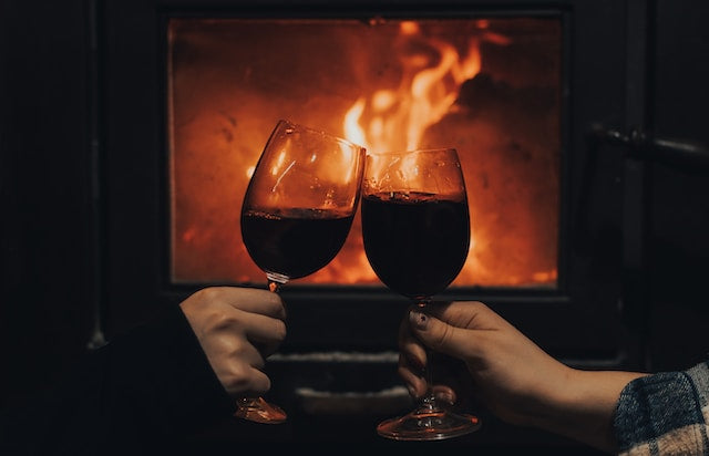 deux personnes trinquant avec leur verre de vin rouge devant un feu de cheminée