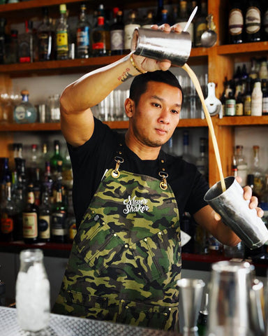 A bartender pours a drink while wearing a camo-coloured luxury bartender apron featuring his custom branded logo.