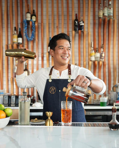 A bartender shakes a drink and pours another while wearing a luxury bartender apron made of denim and featuring a custom embroidered logo.