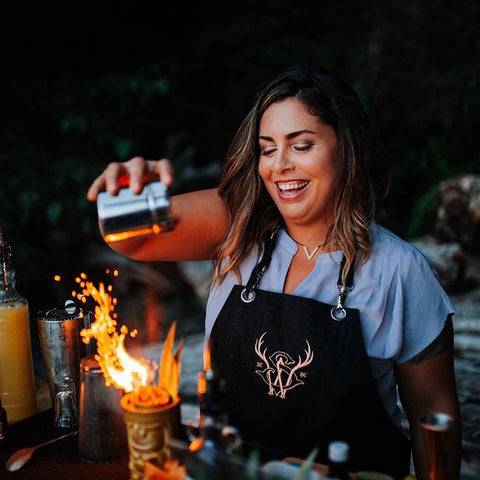 A bartender pours a fiery drink while wearing a luxury bartender apron made of black canvas. The high-end bartender apron features a custom embroidered logo.