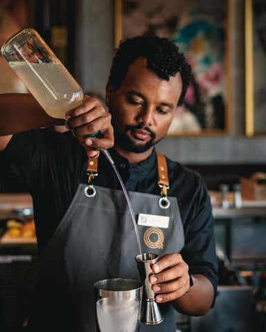 A bartender measures out a shot while mixing a drink. He is wearing a gray canvas luxury bartender apron from S&R Denim Co.