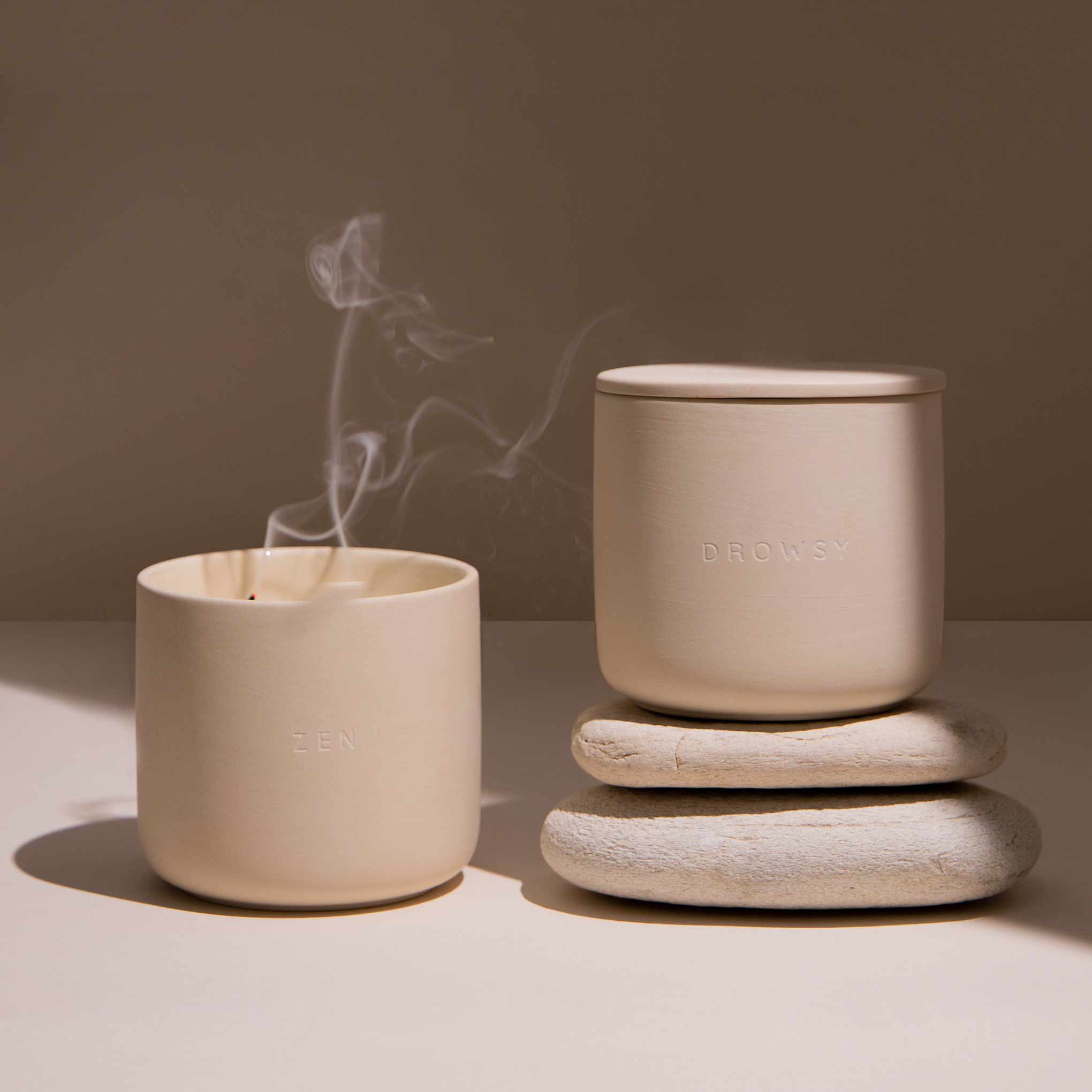 2 Candles in ceramic jars on a cream coloured background. One of them has just been blown out and has smoke gently rising from the wick.