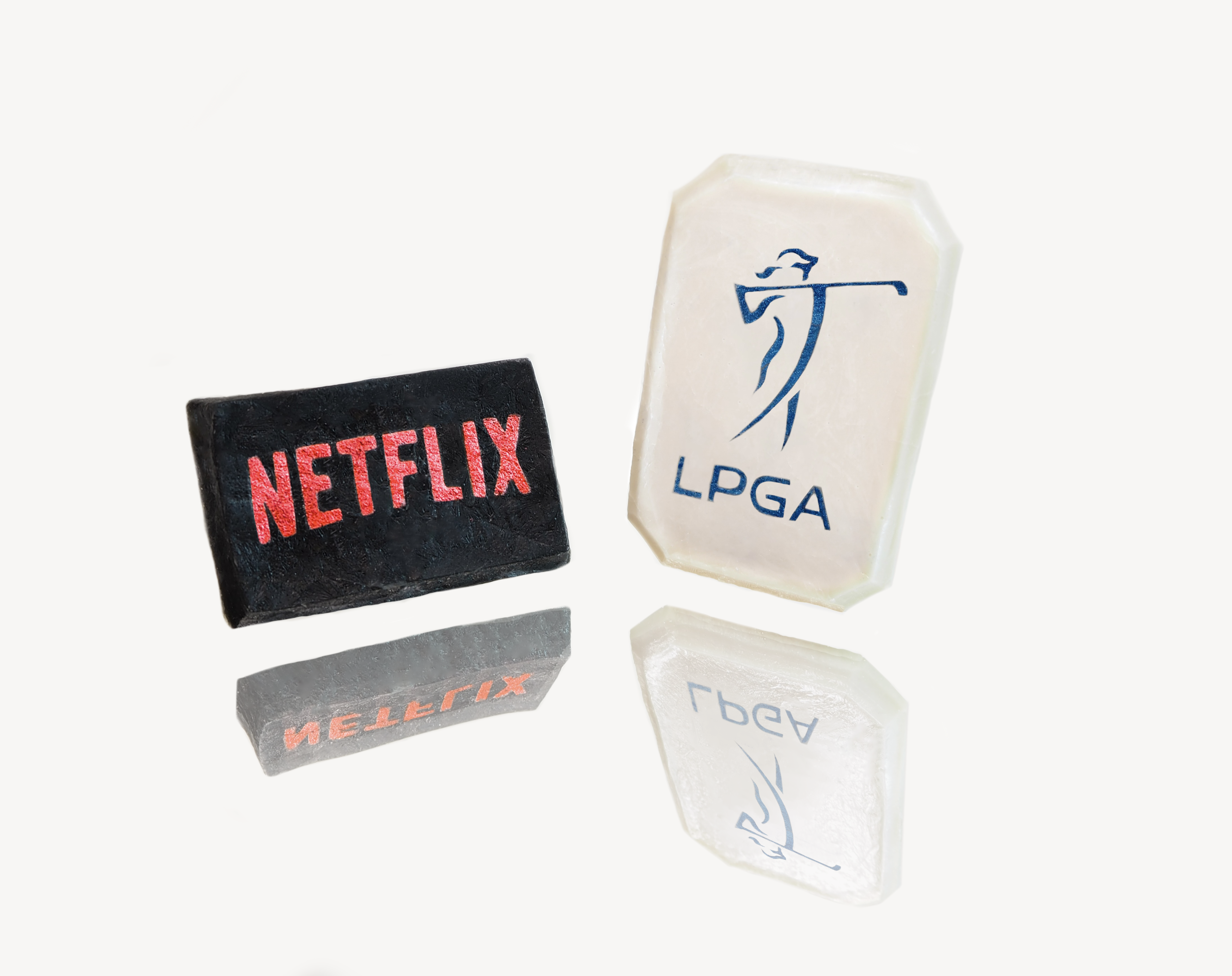 Two fabric patches and two novelty golf-themed sweets designed to resemble soap bars on a reflective surface. The black patch on the left features the 'NETFLIX' logo in red, and the sweets on the right are shaped like soap bars with the 'LPGA' logo in blue, with their reflections clearly visible beneath them