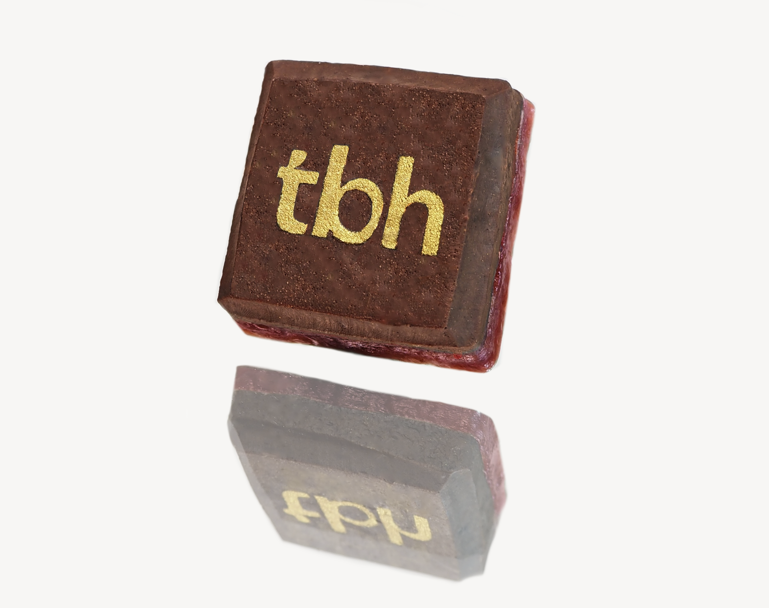 A square chocolate sweet with the letters 'tbh' in gold on top, placed on a reflective surface creating a clear mirror image below it.