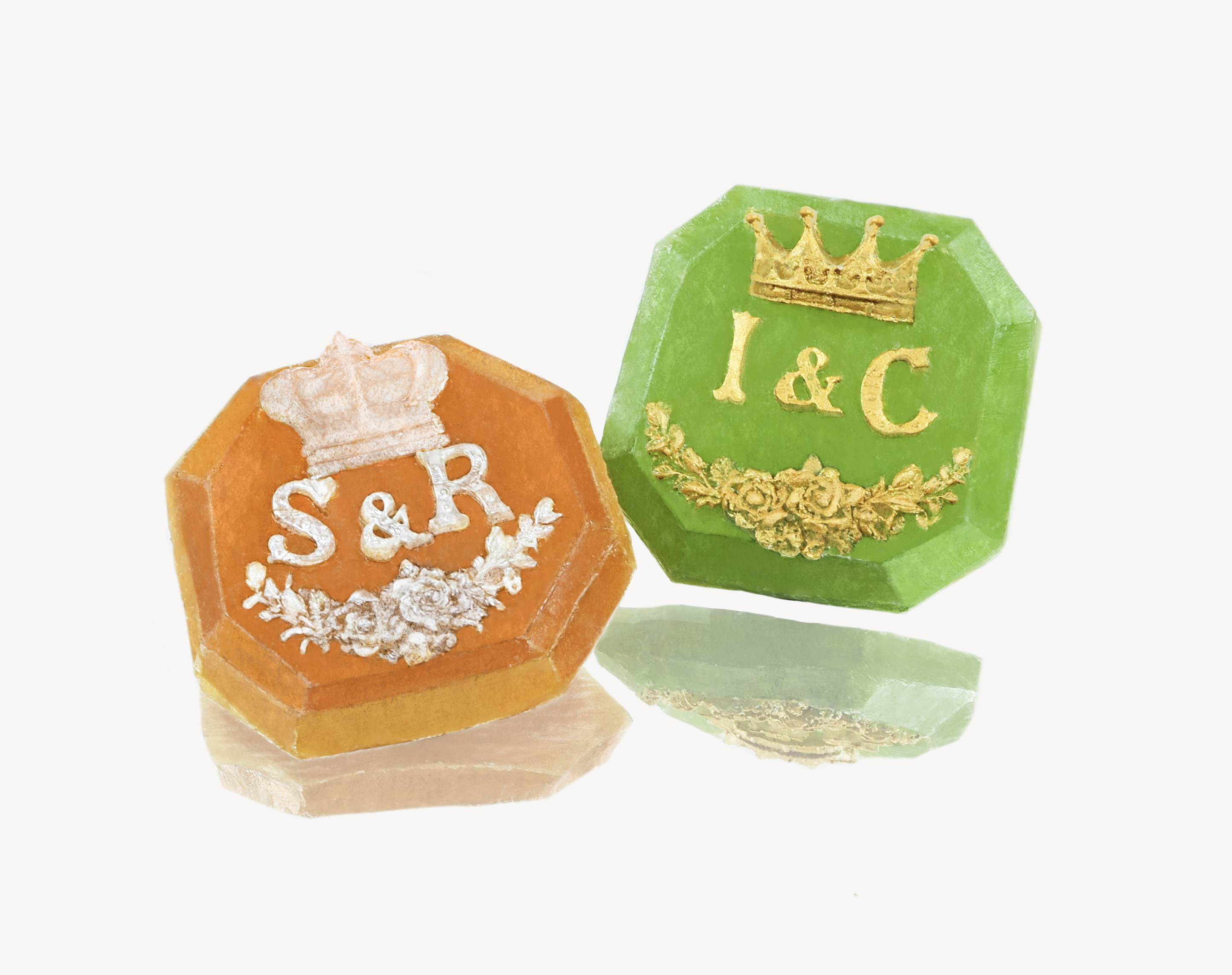 Two octagonal-shaped sweets, one orange with 'S&R' written under a crown and floral design, and the other green with 'I&C' under a crown and surrounded by a floral wreath, both with a gold decorative finish. They are placed on a white background with their reflections partially visible.