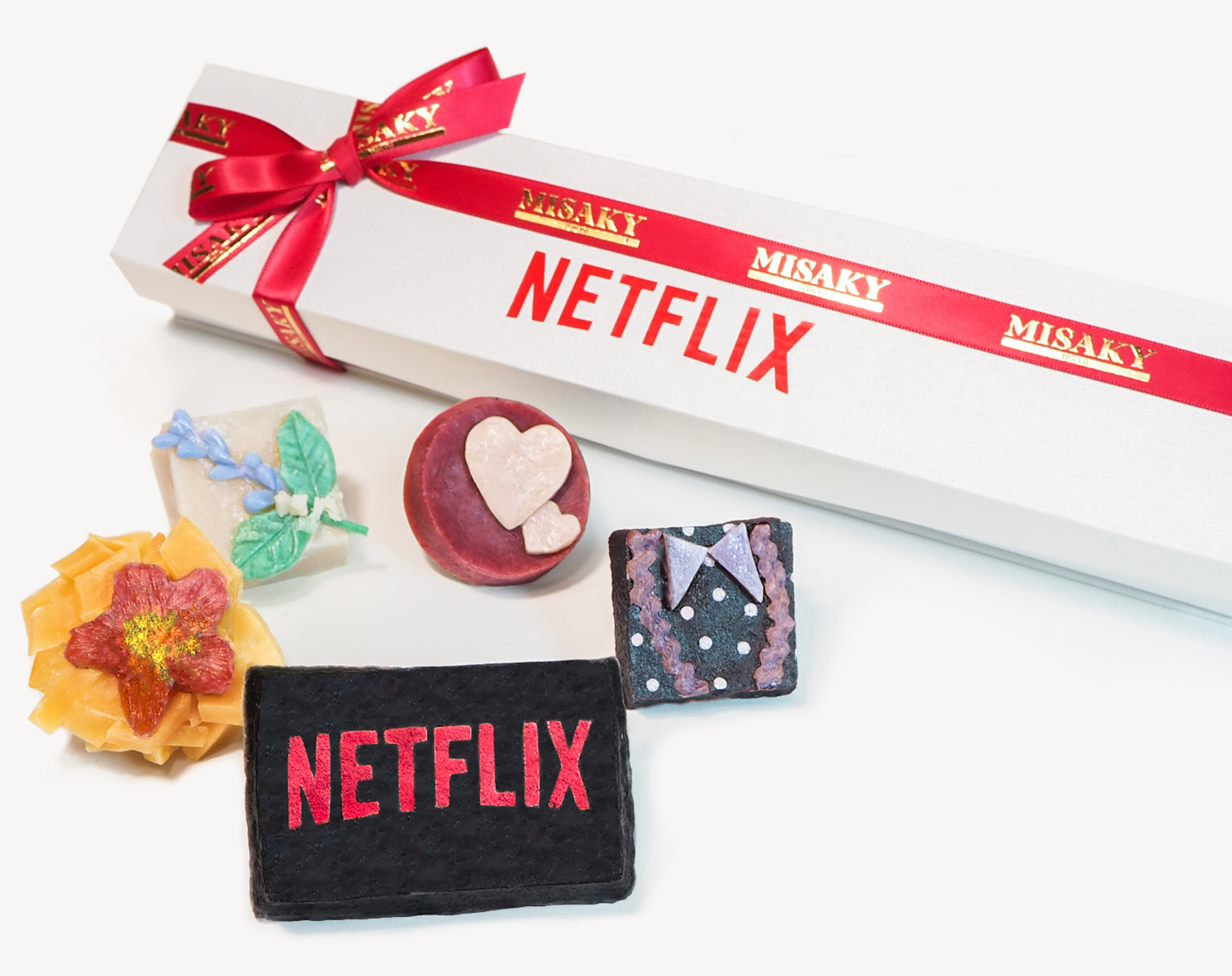 A long white gift box featuring the word 'NETFLIX' in bold black letters, adorned with a red ribbon that has 'MISAKY' printed in gold. Accompanied by decorative items including a red candle with a heart shape, a blue flower, and two fabric patches with 'NETFLIX' logo, set against a white background.