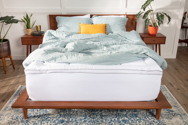 Photo of a white pillow top mattress topper secured to a mattress with a white elastic skirt. There's a pale blue comforter draped across the top.