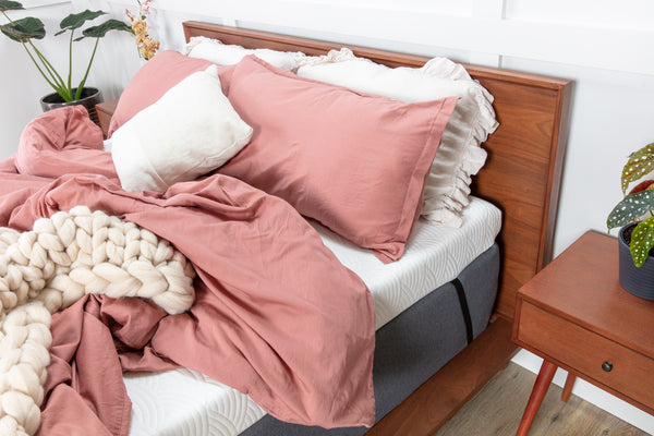 Photo of a thick white mattress topper secured to a mattress with a black adjustable elastic strap. Fluffy rose colored bedding is draped across the top