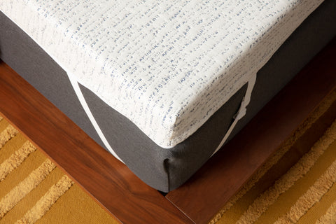 Photo of the corner of a bed with a view from above. The bed has a thick white and blue mattress topper that is attached to the mattress with a white elastic adjustable strap