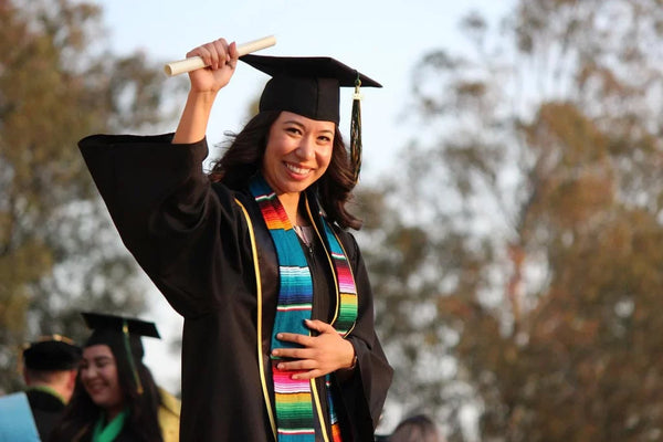 Woman in graduation cap and gown, holding up her diploma and smiling.