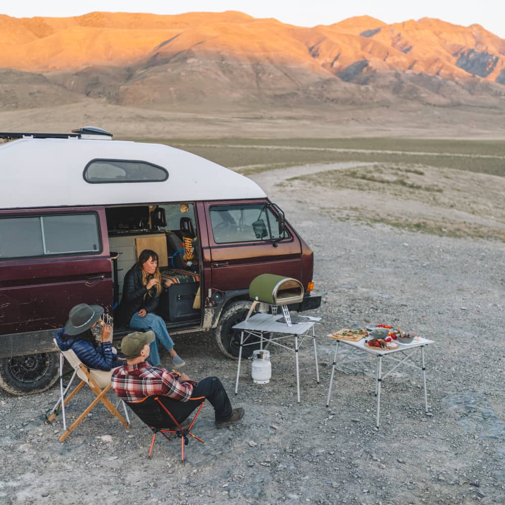 A high vantage shot of a group of friends sitting round and relaxing next to their van as a pizza cooks in the Gozney Roccbox pizza oven