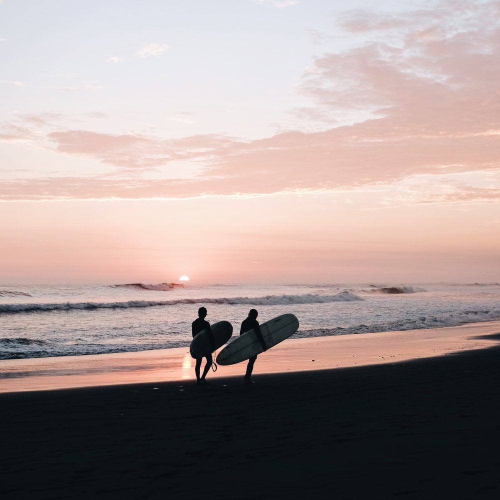 Two surfers stroll along a beach as the surf breaks and the sun sets in the background.