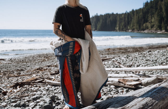 a woman stands on a beach in the PNW wrapped in a nomadic blanket