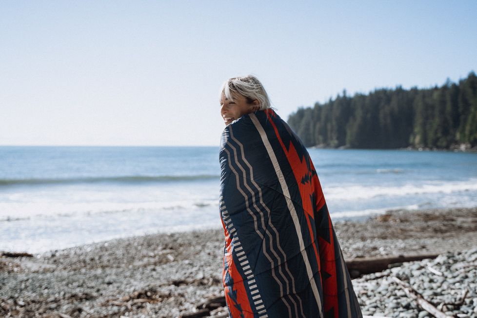 A woman wraps herself in a 2 person Nomadic blanket on a beach surrounded by trees