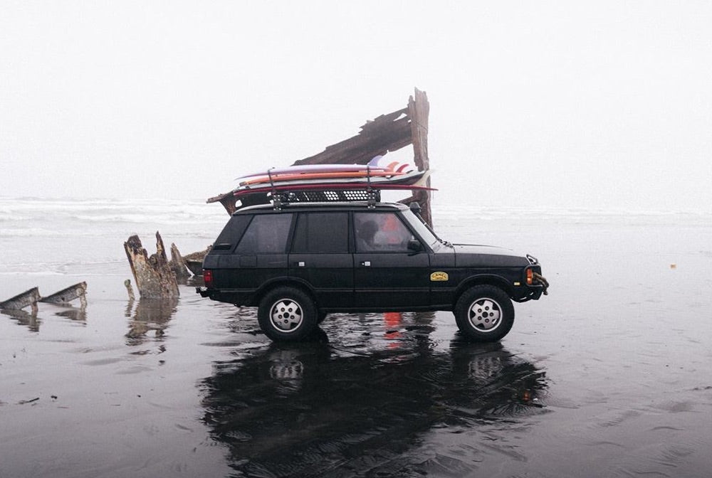 A 4x4 sits parked on a beach next to a giant piece of drift wood