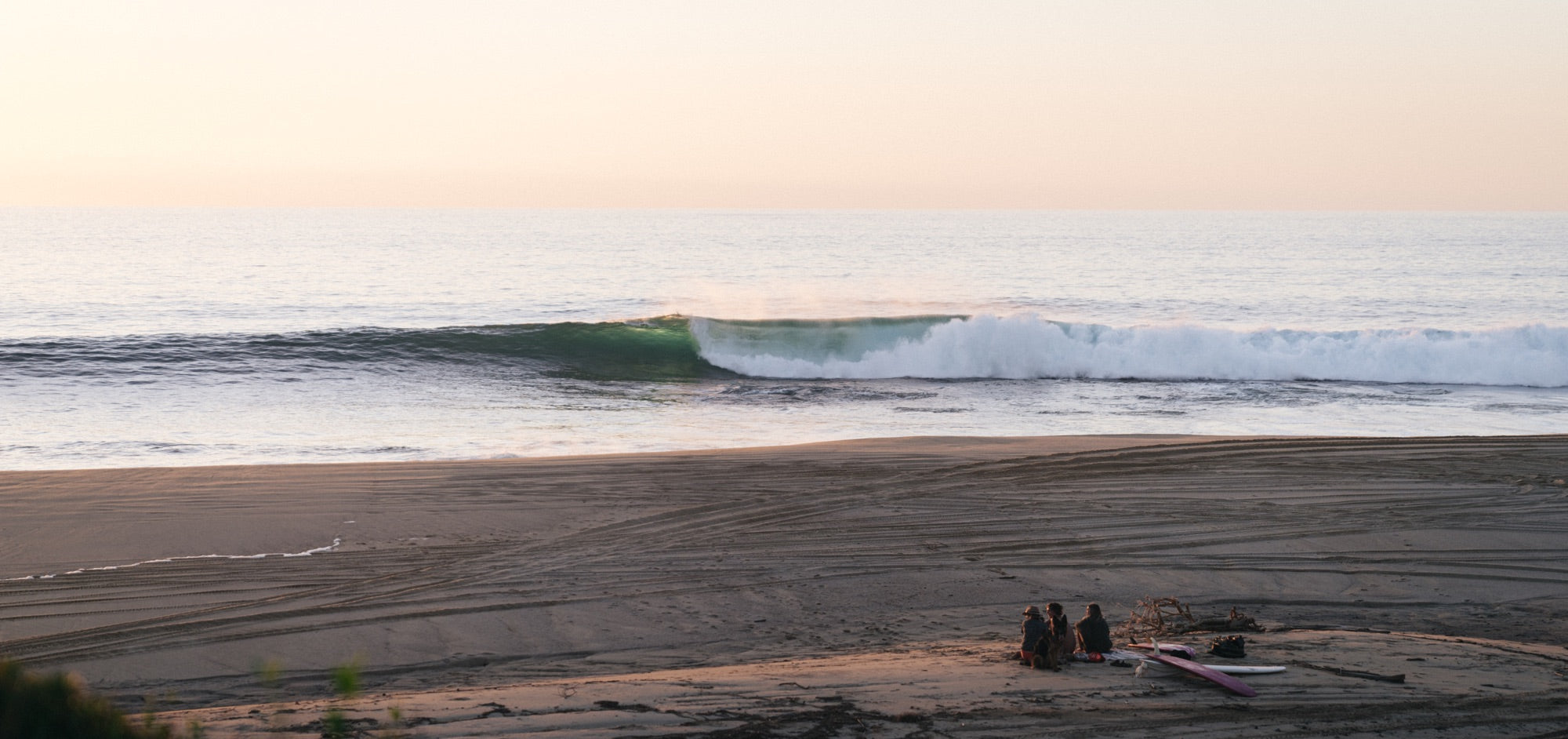 A group of friends sit on the beach at dusk as a perfect wave rolls through