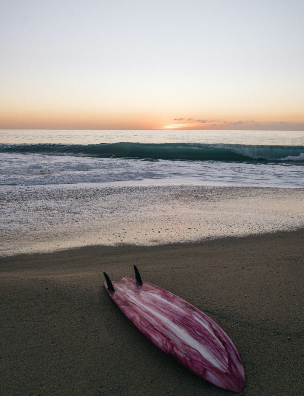 A twin fin surfboard lays in the sand at sunset as a wave breaks in the background