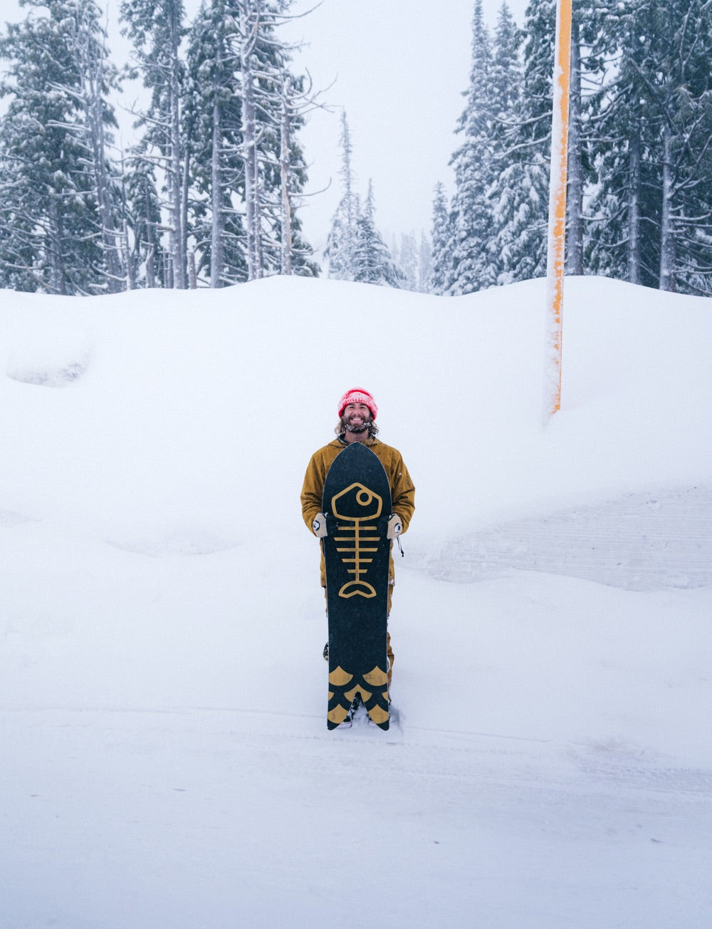 Brandon Haley stands on the side of a road holding his snowboard and a big grin, with a snowy forest scene in the background