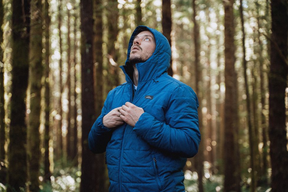 A man stands in a forest looking up at the trees wearing a patrol jacket