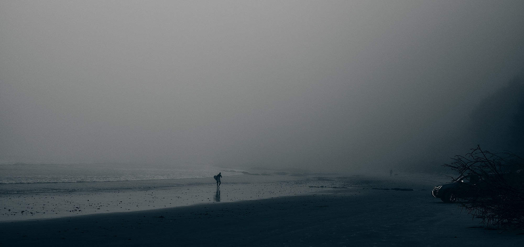 A surfer walks along a beach shrouded in mist as another figure approaches from the distance