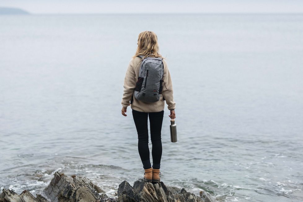 A woman stands on rocks looking out to sea wearing the vale backpack