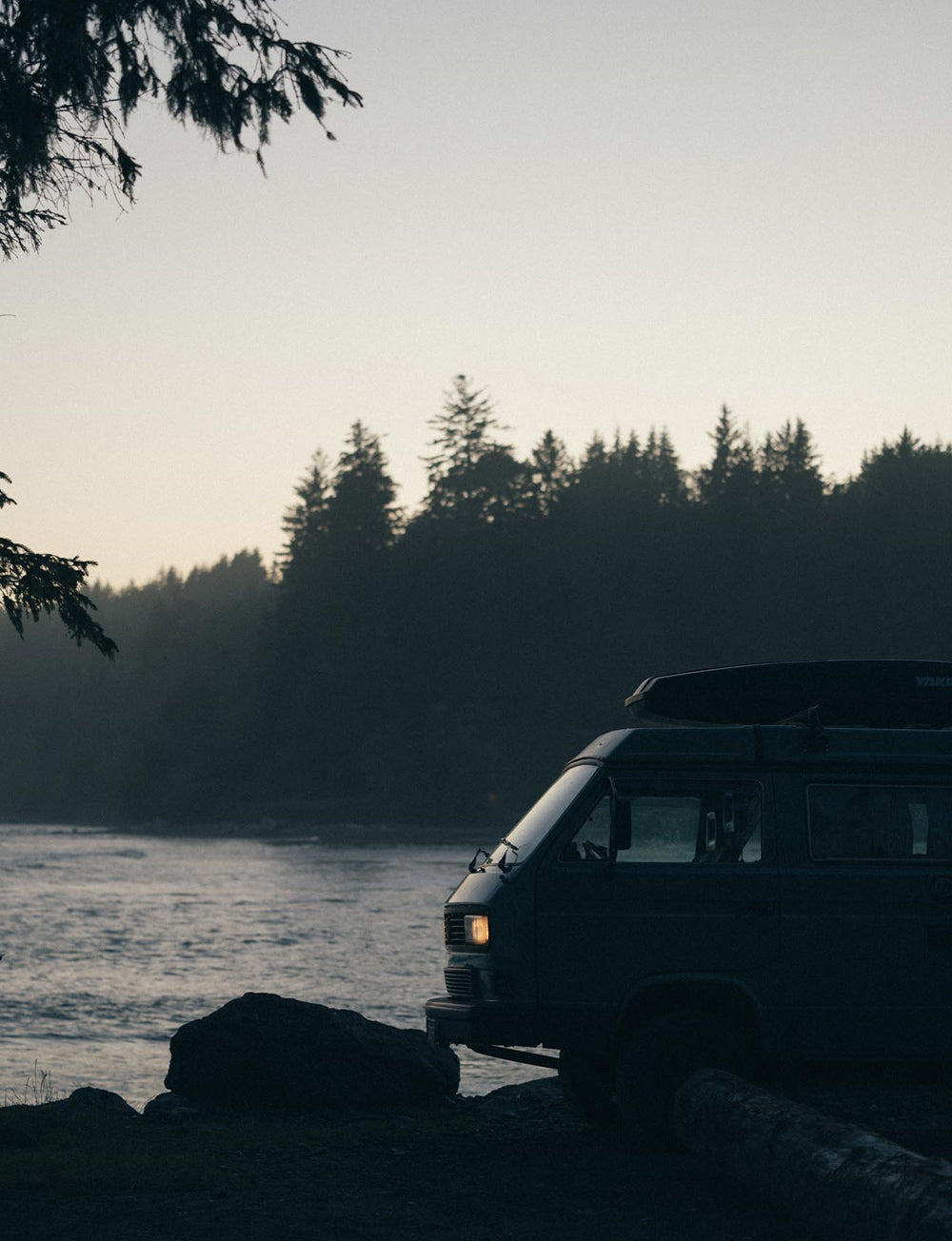 A classic VW Westy sits by the water during blue hour with trees and ocean in the background.