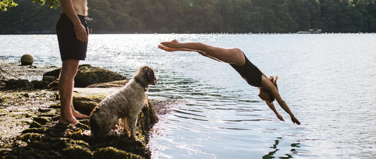 A woman dives into a lake while a man and his dog watch from the shore