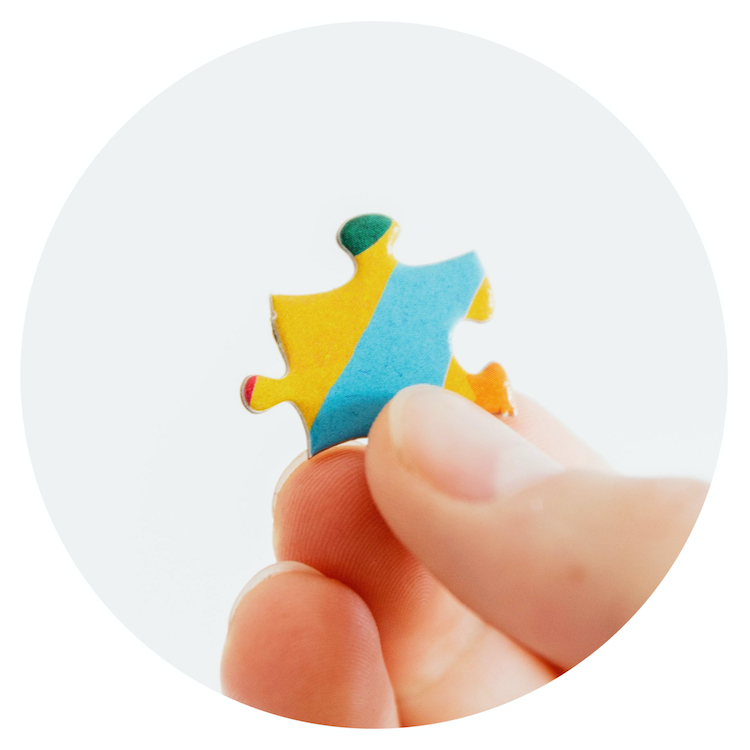 a small round image of a caucasian hand holding up a yellow, teal, and green puzzle piece in front of a grey background.