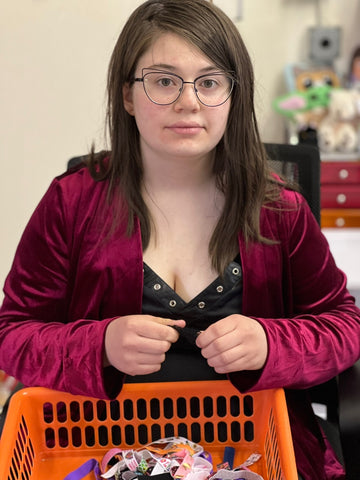 A teen girl with brown hair and cat eye glasses in a red coat. She's working on making ponytail holders that she has in an orange plastic bin in front of her.