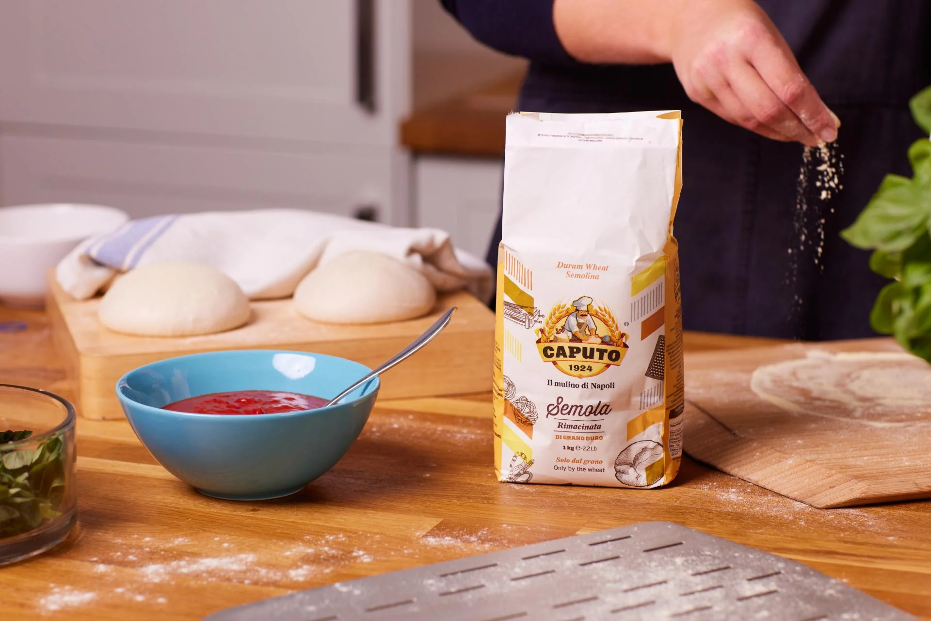 One hand sprinkling Caputo Semola fFour over an Ooni Bamboo Pizza Peel & Serving Board on a table with a bag of  Caputo Semola Flour, a bowl of tomato sauce and Ooni dough balls.