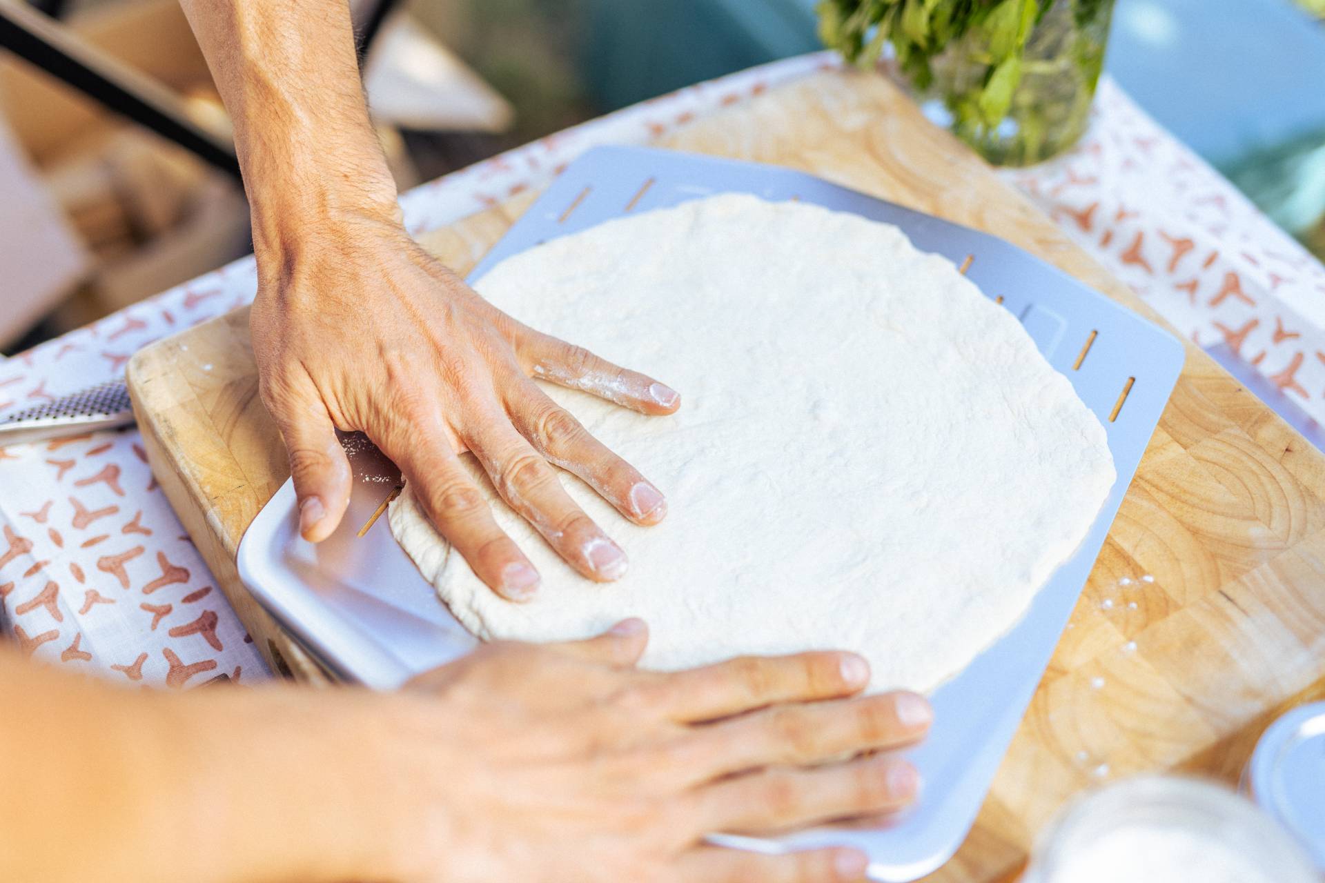 Two hands stretching dough on an Ooni Perforated Pizza Peel on top of a bamboo cutting board.