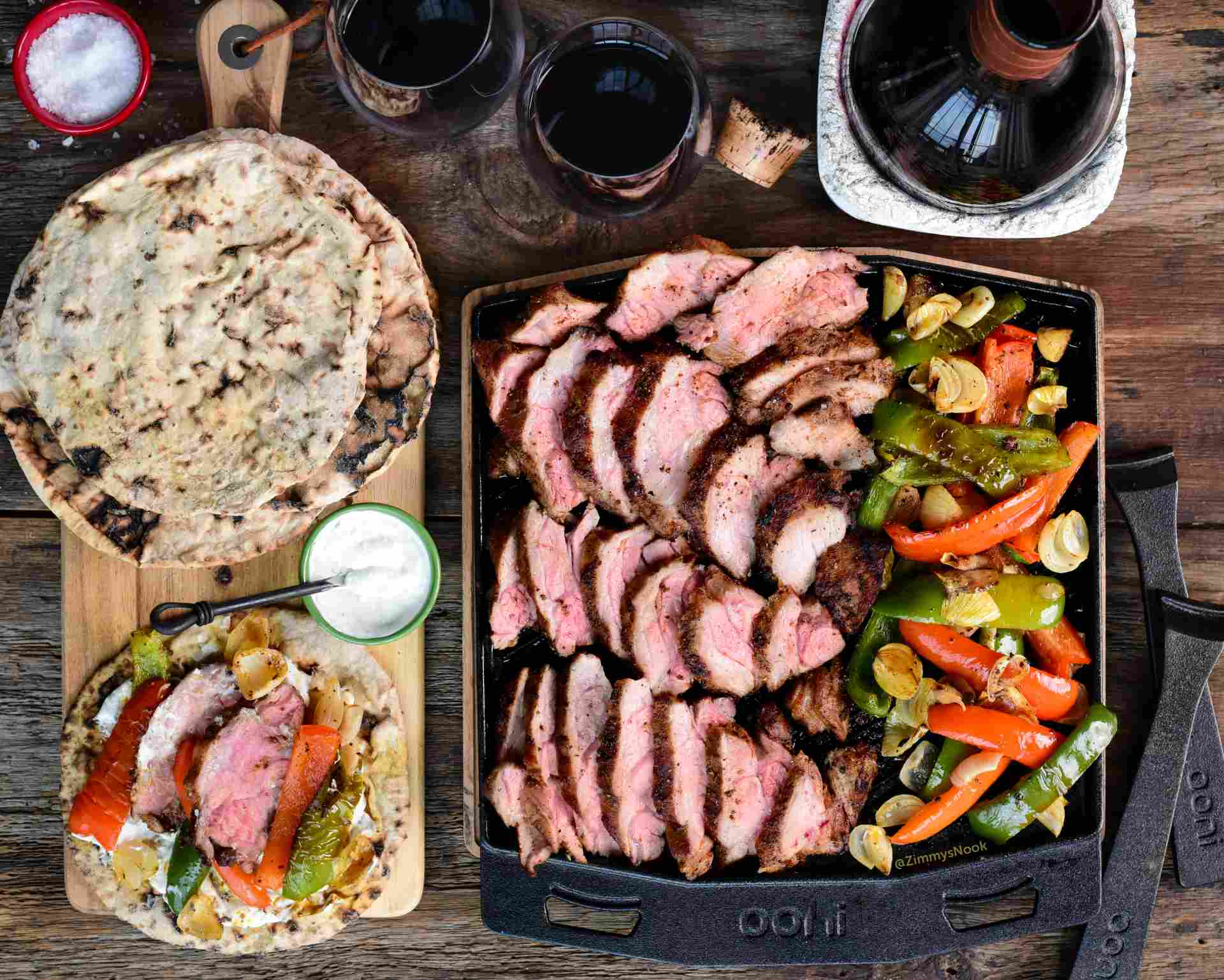 Slices of grilled harissa spiced lamb neck, red and green bell peppers and onions on an Ooni Dual-Sided Grizzler Plate next to a wooden serving board with flatbreads and tahini sauce on a table.