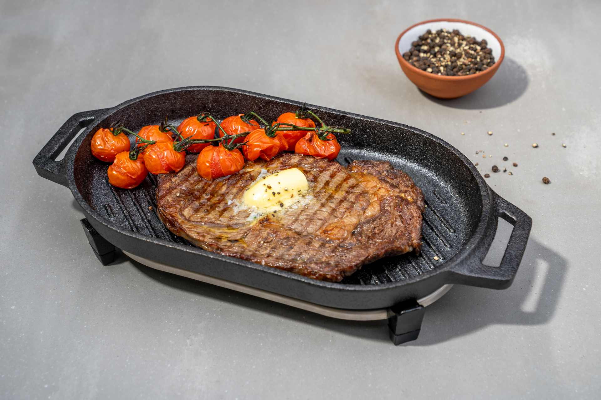 Cooked steak with butter and roasted tomatoes in an Ooni Cast Iron Grizzler Pan on a table.