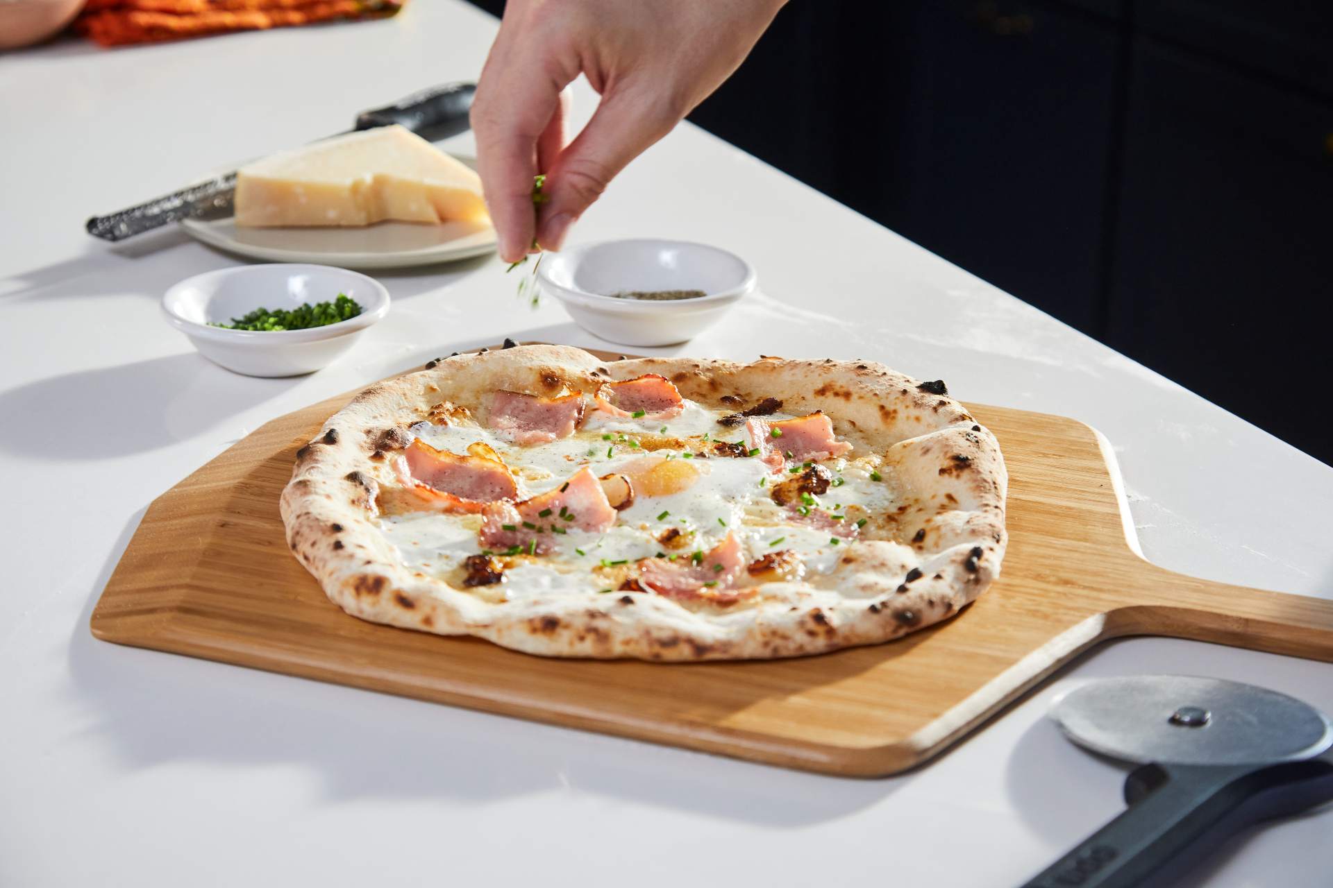 A pizza topped with bacon, eggs and cheese being topped with fresh herbs