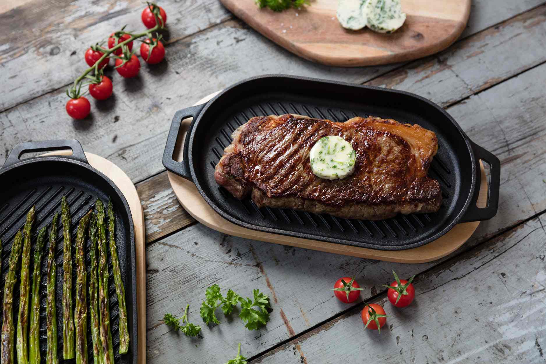 Grilled steak with a pat of butter on an Ooni Cast Iron Grizzler Pan next to grilled asparagus on an Ooni Cast Iron Grizzler Pan on a table.