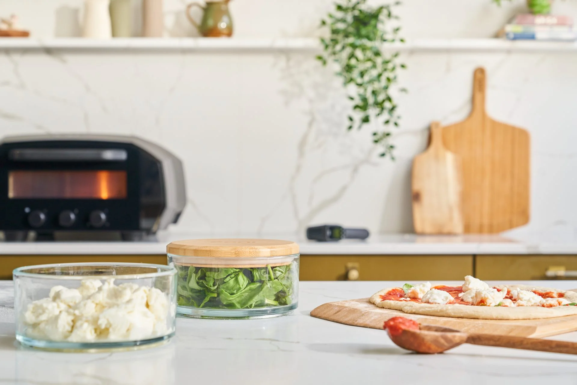 Ooni Stack with fresh mozzarella and basil on table with an uncooked margarita pizza on an Ooni Bamboo Pizza Peel & Serving Board with a Volt 12 in the background.